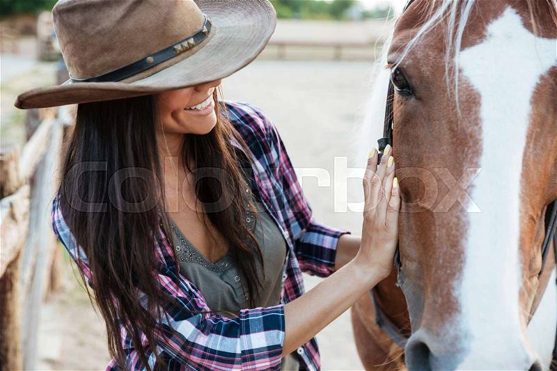 Closeup of smiling tender young woman cowgirl in hat with her horse, stock photo