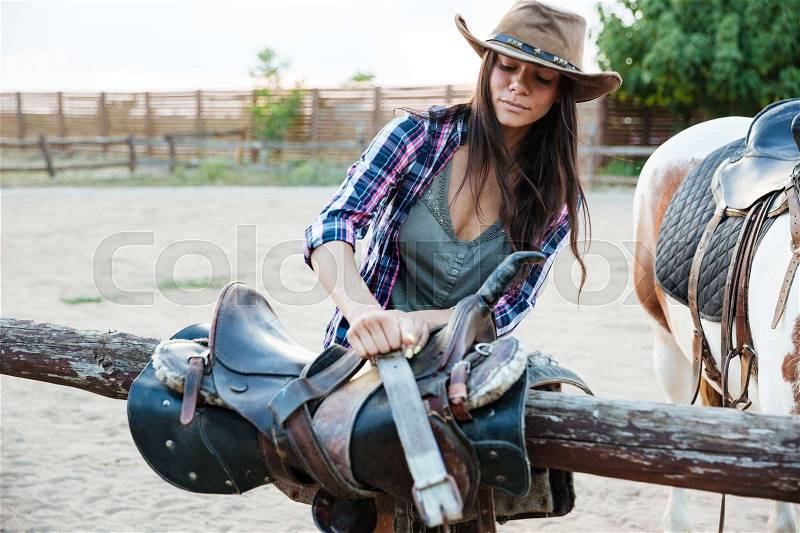 Beautiful young woman cowgirl in hat preparing saddle for riding horse, stock photo