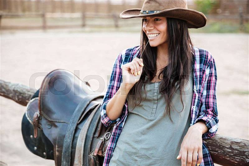 Happy smiling cowgirl in hat holding straw in mouth while leaning on the ranch fence, stock photo