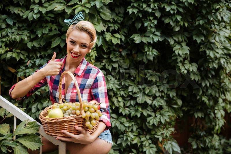 Cheerful pinup girl with basket of fruits showing thumbs up, stock photo