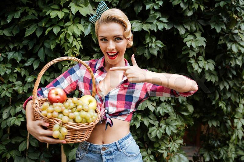 Cheerful pin up girl pointing on basket of fruits, stock photo
