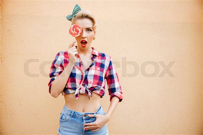 Surprised cute pinup girl covered her eye by lollipop, stock photo