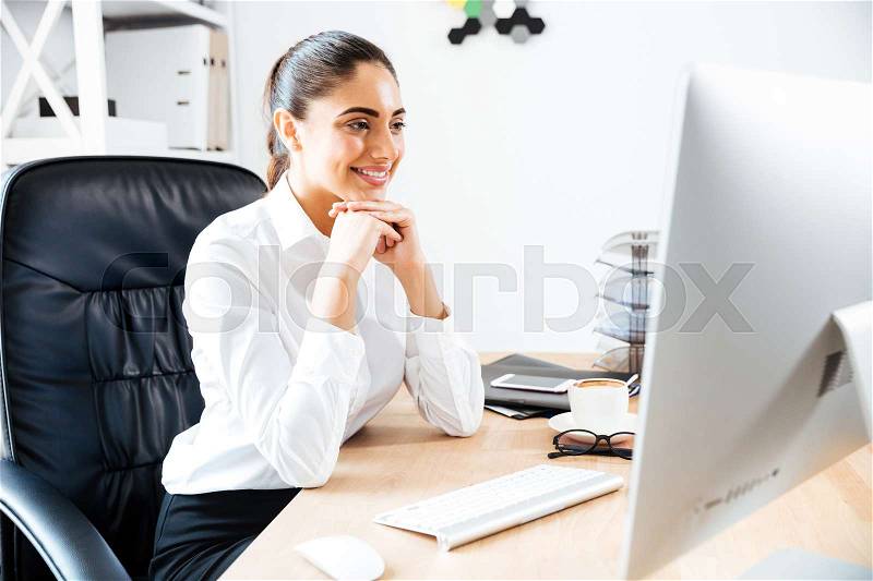 Happy young businesswoman looking at computer screen while sitting at the office table, stock photo