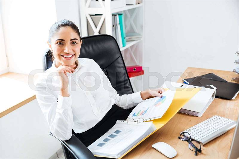 Smiling happy businesswoman holding reports and looking at camera while sitting at the workplace, stock photo