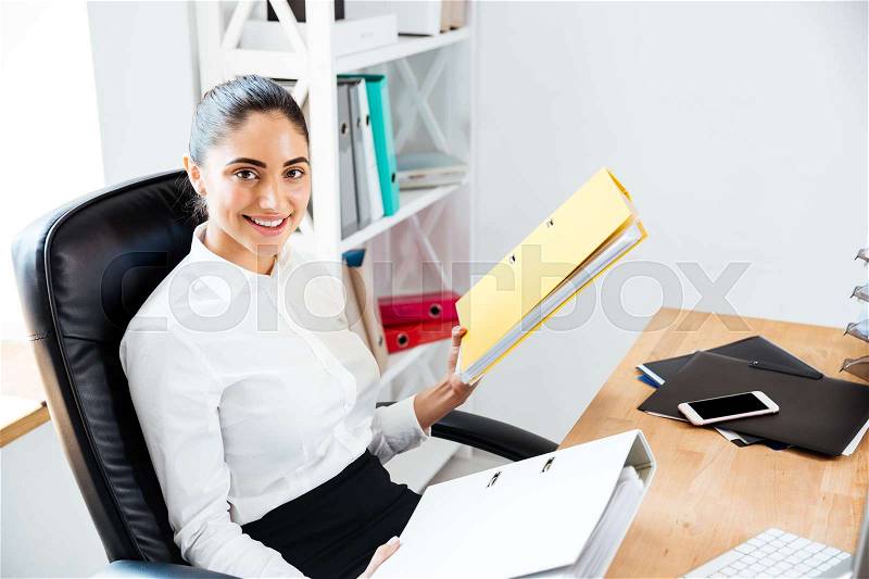 Happy beautiful businesswoman holding binders and looking at camera while sitting at the office, stock photo