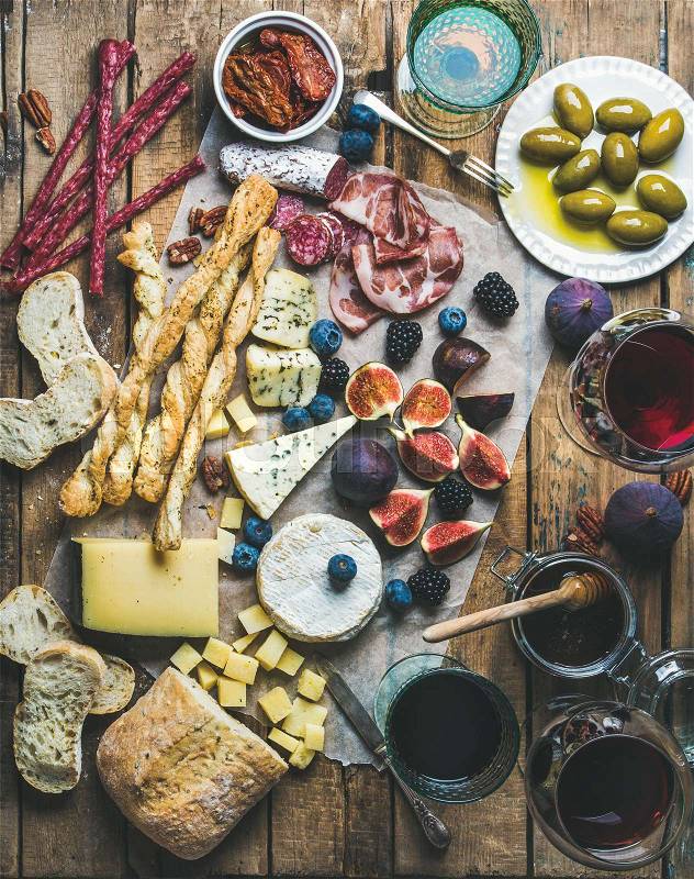 Wine and snack set with various wines in glasses, meat variety, bread, green olives, sun-dried tomatoes, figs, nuts and berries on wax paper over rustic wooden background, top view, stock photo