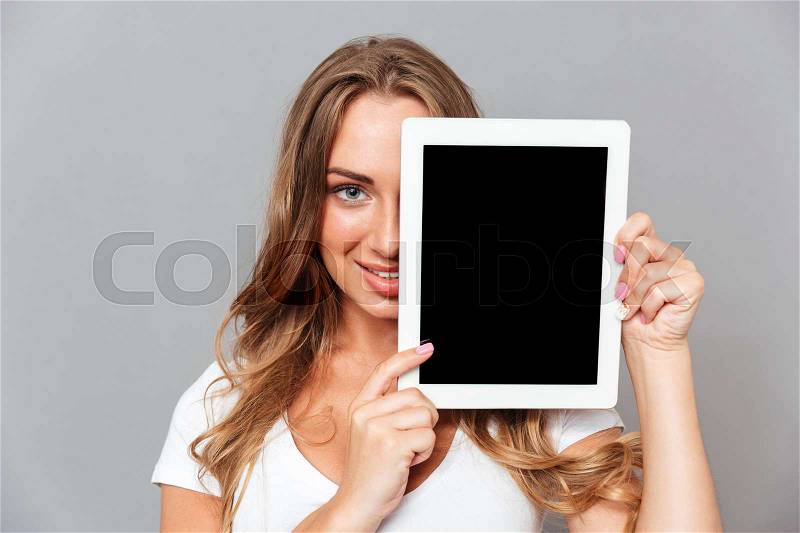 Smiling young woman covers her face with blank screen tablet computer isolated on a gray background, stock photo