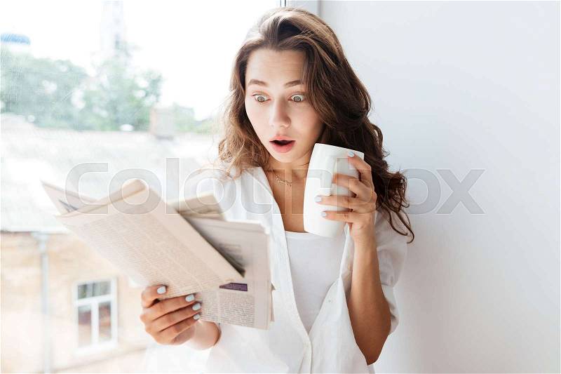 Surprised young brunette woman looking at newspaper and holding tea cup while sitting on the windowsill, stock photo