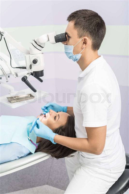 Nice girl with patient bib on a dental chair and dentist who sits next to her. He looks on her teeth using dental microscope and holds dental bur and mirror. Vertical, stock photo