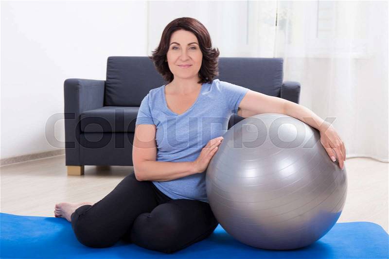 Beautiful slim sporty mature woman with fitness ball at home, stock photo