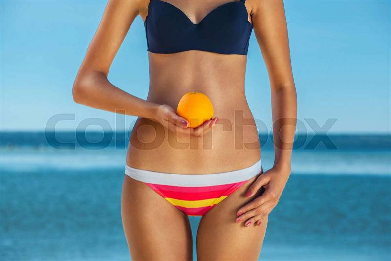 Young, skinny, pretty girl in a striped bathing suit holding a orange on the beach and the sea. The concept of healthy nutrition and body care, stock photo