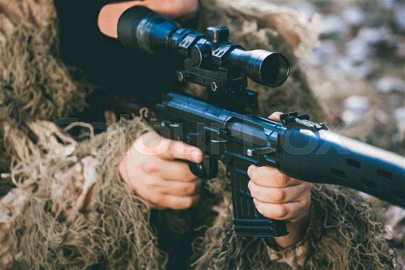 The soldier disguised in a balaclava and military camouflage aims at the sight of a sniper rifle. War Zone, stock photo