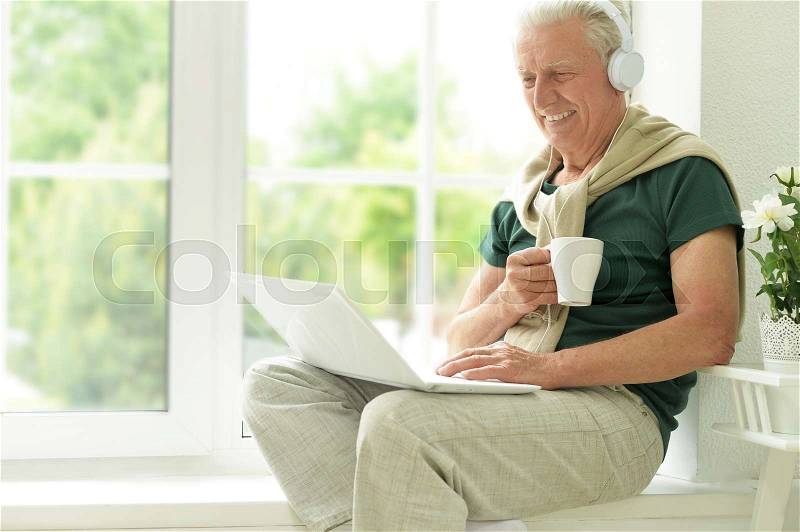 Elder man with laptop and headset sitting on the windowsill at home, stock photo