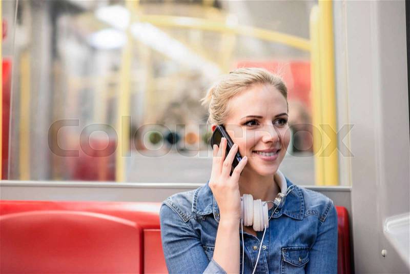 Beautiful young blond woman in denim shirt in subway train, holding a smart phone, making a phone call, stock photo