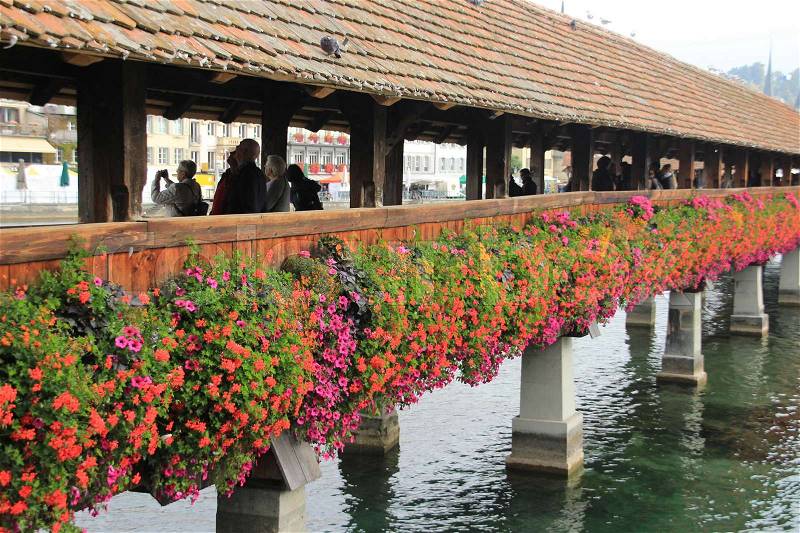 Many people walk over the wooden bridge, called Kapellbrücke with blooming geranium and petunia in different colours in the city Lucerne in Switzerland in fall, stock photo