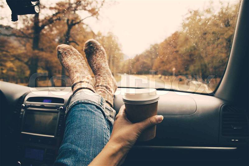 Woman feet in warm socks on car dashboard. Drinking take away coffee on road. Fall trip. Rain drops on windshield. Freedom travel concept. Autumn weekend. Filtered photo, stock photo