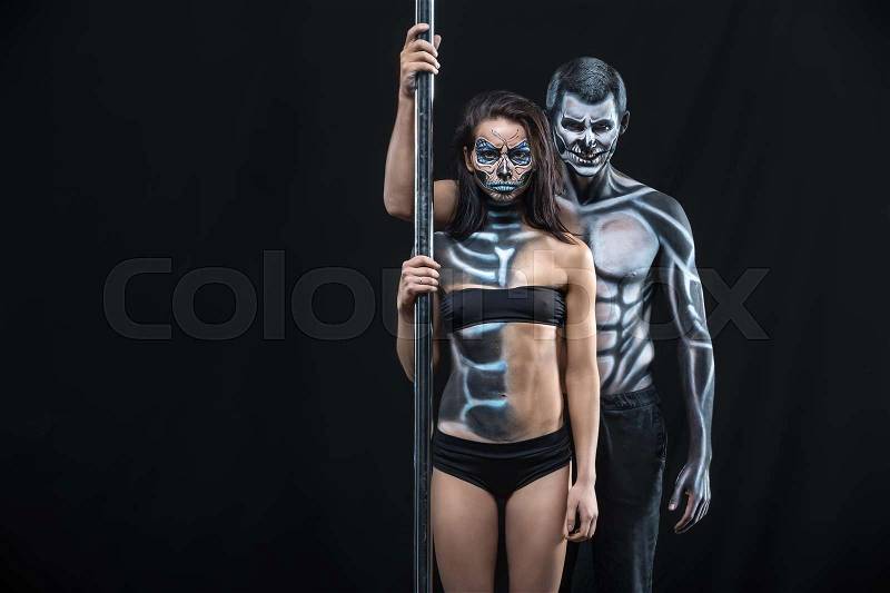 Grinning couple of pole dancers with horrific body-art stands next to a pylon on a dark background in the studio. They dressed in black sportswear and hold the right hands on the pylon. Horizontal. , stock photo