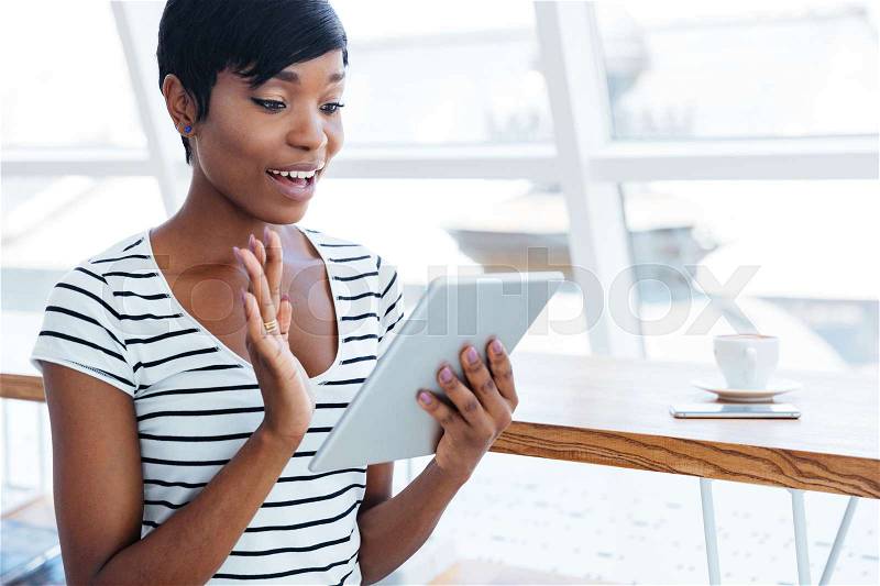 Happy afro american businesswoman holding tablet computer and waving in office, stock photo