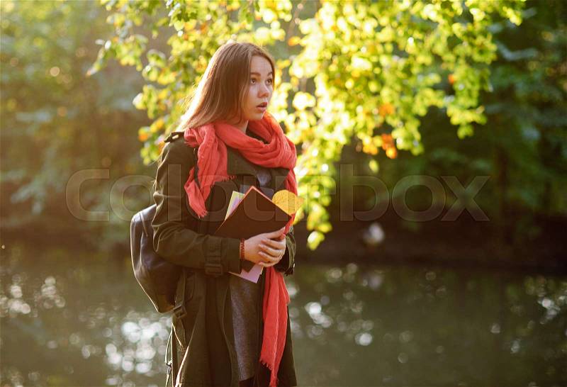 Autumn. Lovely girl in a bright red scarf walks in autumn park. Trees reflected in the pond. Through foliage the sunlight makes the way. In the hands of a girl is notebooks, backpack on her shoulder, stock photo