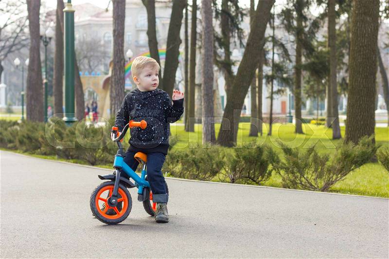 Little boy on the run bike is standing and raising one hand, stock photo