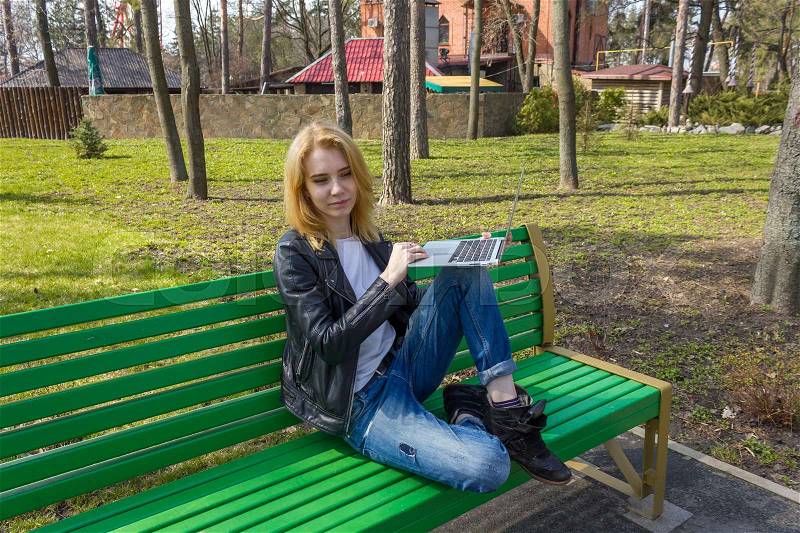 Woman sitting with one leg up on bench and holding silver laptop, stock photo