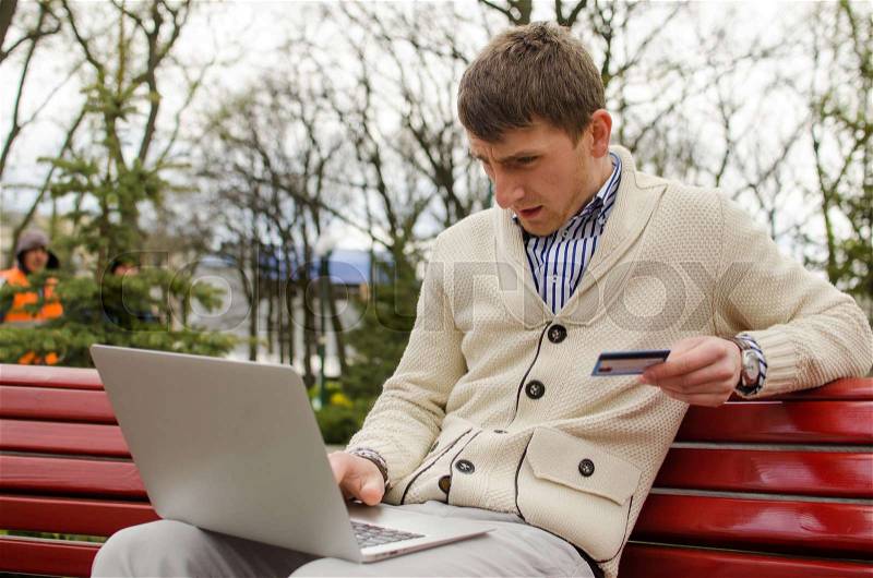Young man with laptop in park in white pullover, stock photo