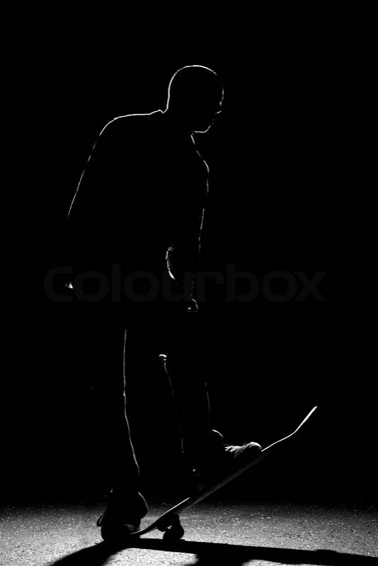 A backlit skateboarder guy posing under dramatic rim lighting with his skateboard flipped up in the front, stock photo