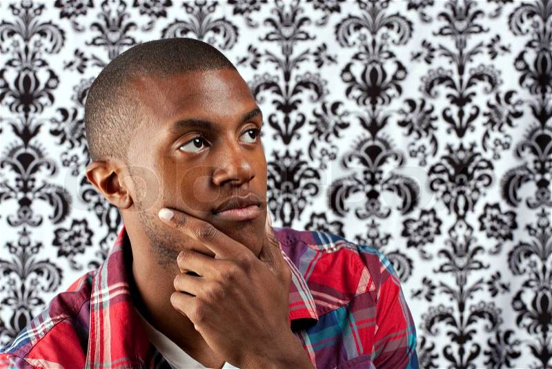 A young African American man in his twenties and his hand on his chin thinking deeply about something in front of a damask style background. Shallow depth of field, stock photo