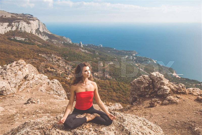 Young pretty woman is sitting in yoga pose asana in beautiful sea and mountains landscape view from the cliff, stock photo