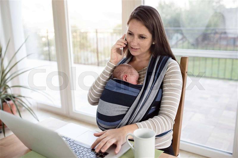 Beautiful young mother with her newborn baby son in sling at home, sitting at the table, writing on notebook keyboard, holding smart phone, making phone call, stock photo