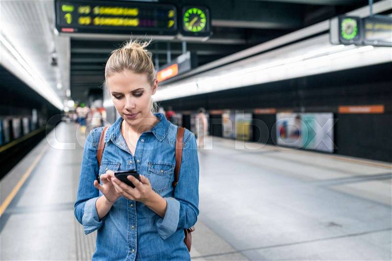 Beautiful young woman in denim shirt with smart phone, standing at the underground platform, texting, stock photo
