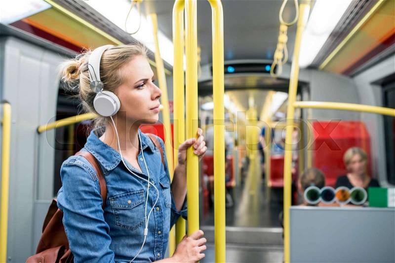 Beautiful young blond woman in denim shirt standing in subway train with white headphones listening music, stock photo