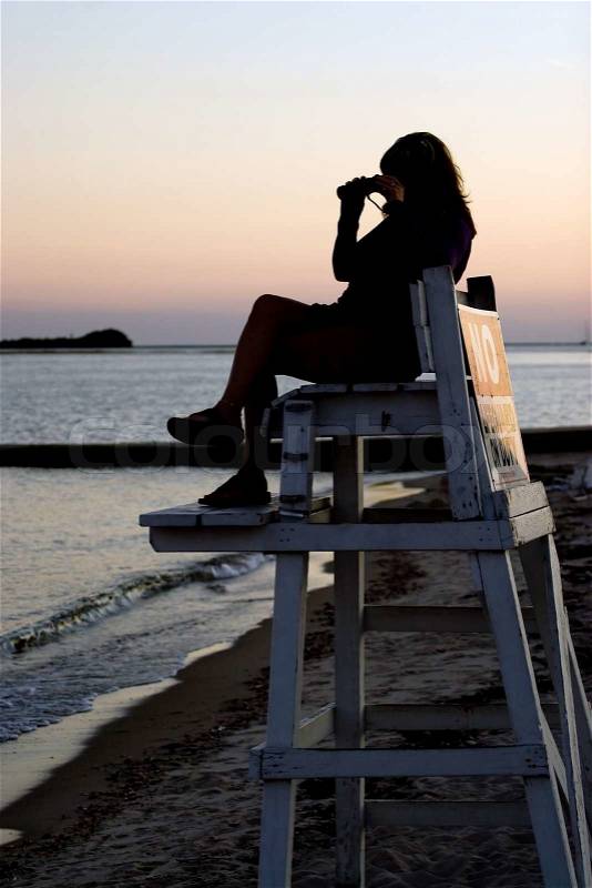 A silhouette of a woman looking with binoculars at the beach while seated in a lifeguard chair, stock photo