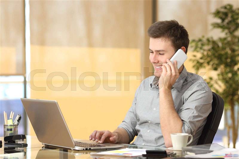 Freelance working on line and talking on phone while is searching information in a laptop sitting in a desk at office, stock photo