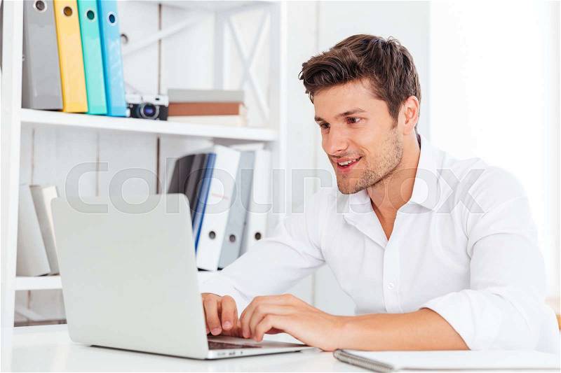 Smiling young happy businessman using laptop computer and sitting in office, stock photo
