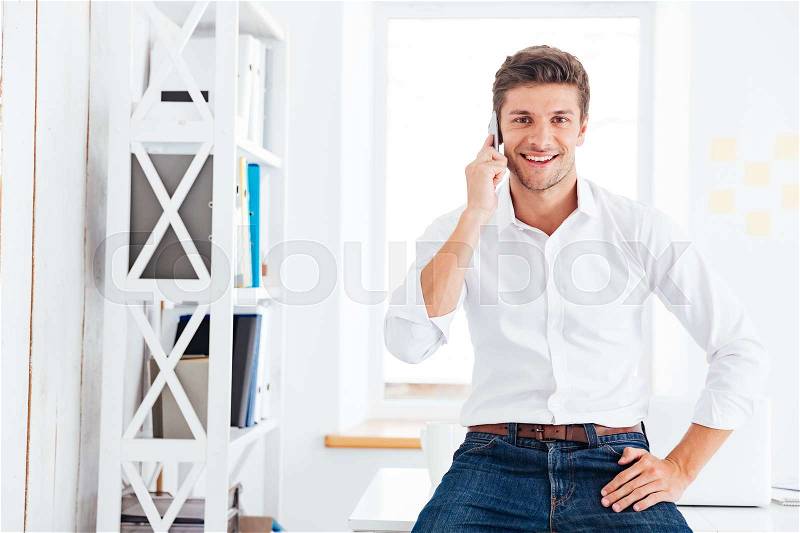 Happy businessman sitting on the table and talking on the phone in office, stock photo