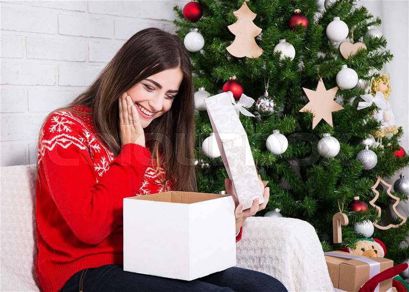 Young happy surprised woman opening gift box near decorated christmas tree, stock photo