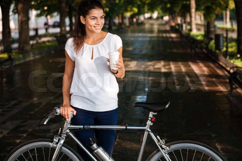 Cheerful brunette woman with takeaway coffee cup and bicycle in the park, stock photo
