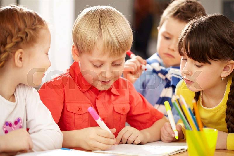 A little boy sitting at table and drawing and his friends looking at him, stock photo