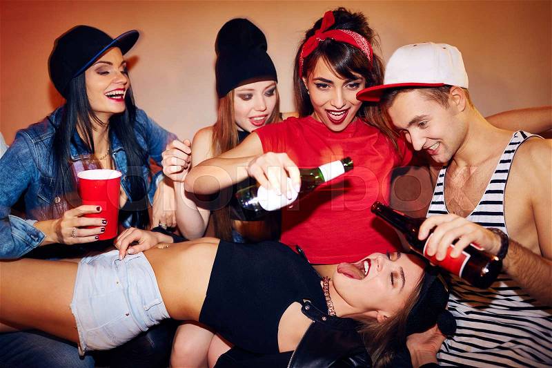 Group of friends crashing at noisy students house party, they pour beer from bottles right into mouth of sexy half naked girl lying in their lap, stock photo