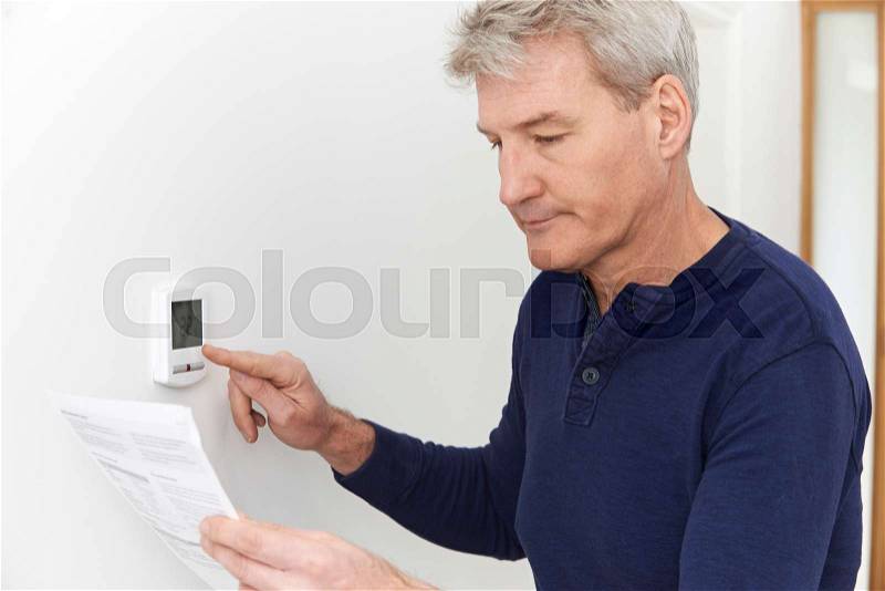 Worried Mature Man With Bill Turning Down Heating Thermostat, stock photo