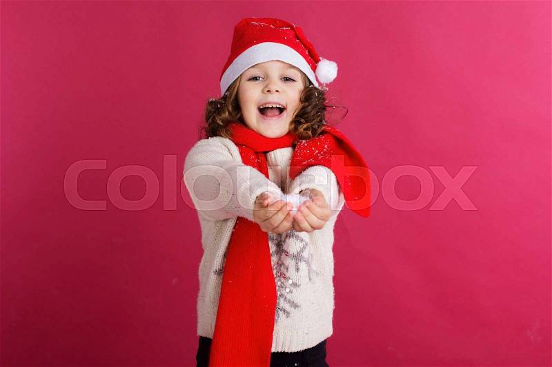 Little cute child girl is holding fake snow in hands wearing warm clothes and santa hat isolated on red background, stock photo