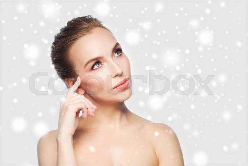 Beauty, people, winter and health concept - beautiful young woman touching her face over gray background and snow, stock photo