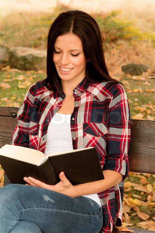 Beautiful young woman reading a book sitting on a bench in park at fall, stock photo