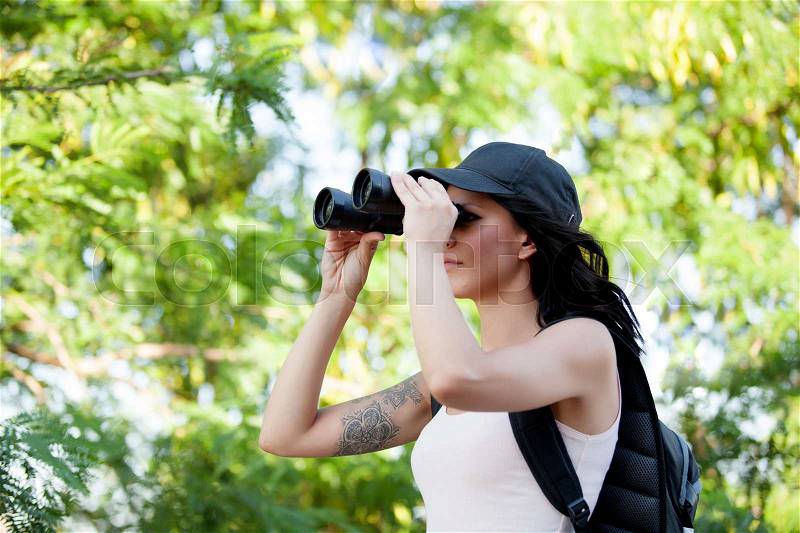 Beautiful woman with cap and backpack looking through binoculars while hiking trip, stock photo
