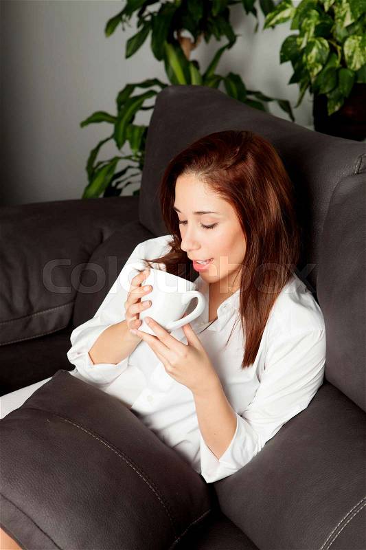 Cute young drinking a hot cup of tea sitting on the couch at home, stock photo