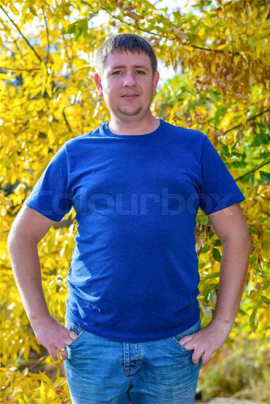 Friendly chubby man standing with his hands in his pockets in front of colorful yellow autumn foliage looking at the camera with a quiet smile, stock photo
