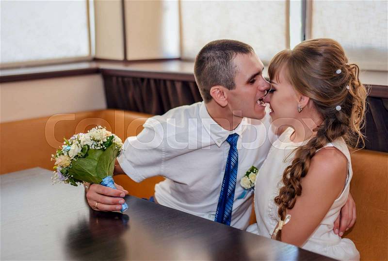 Enthusiastic husband bites the nose of his wife as they sit at a corner booth and while he holds a bouquet, stock photo