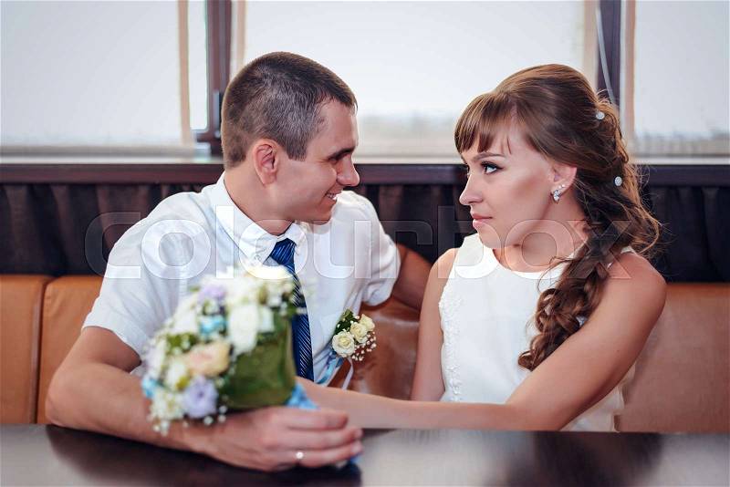 Smiling newlyweds sit at corner booth by windows while holding floral bridal bouquet, stock photo