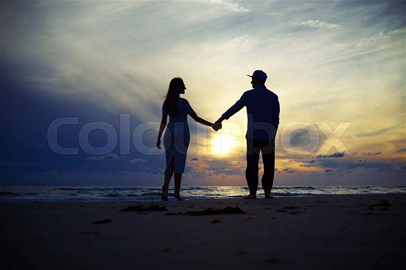 Wide low angle shot of romantic couple holding hands together at the sunset. Young couple holding hands at beach sunset enjoying romance and sun. Young happy couple in love on romantic summer holidays vacation, stock photo